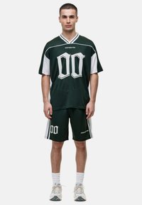 Groene en witte sporttop van mesh-materiaal, met grote witte "00" opdruk. Gecombineerd met shorts die hetzelfde logo tonen.