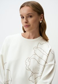 Witte sweatshirt met een oversized pasvorm, voorzien van een grote beige florale omtrek en kleine strasssteentjes aan één kant. Zachte textuur en ronde halslijn.