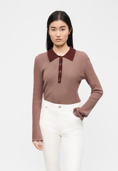 Γυναίκα με μακριά σκούρα μαλλιά που φοράει καφέ μακρυμάνικη μπλούζα με ριπ ύφανση και μπορντό γιακά, καθώς και άσπρο ψηλόμεσο παντελόνι, στέκεται με το ένα χέρι στην τσέπη.