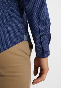 Marinblå skjorta med ett litet geometriskt mönster, knäppt manschett och sidoslitsar, kombinerad med beige byxor som visar en slät textur.