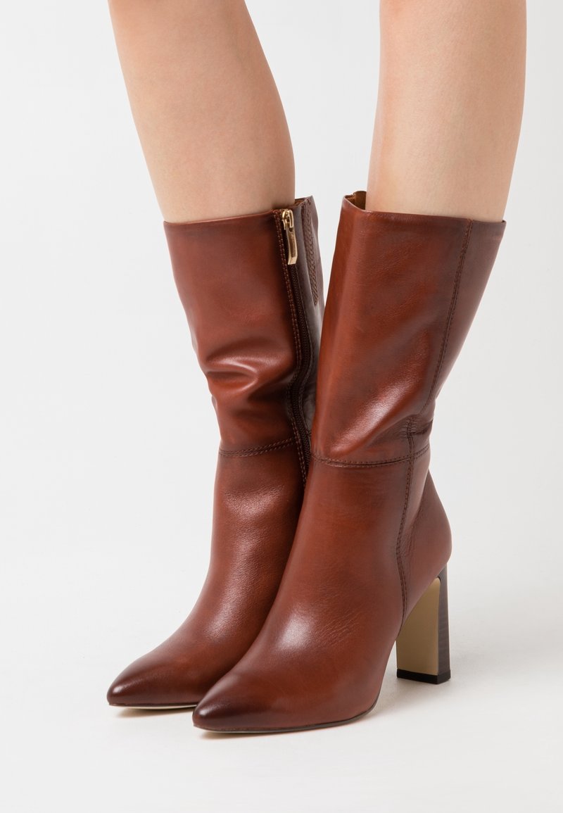 Bottes en cuir marron mi-mollet avec bouts pointus, talons blocs et fermetures éclair latérales portées sur des jambes nues sur un fond uni.