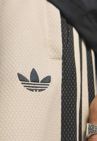 Bežové športové šortky s textúrovanou tkaninou, so zwartými pruhovanými akcentmi a vyšitým čiernym logom na ľavej strane.