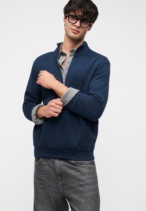 Marineblauwe sweatshirt met rits en een gestructureerde onderste paneel, geribbelde manchetten en een gelaagde grijze overhemdkraag eronder. Draagt een grijze spijkerbroek.