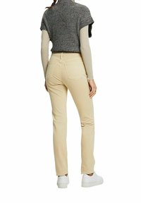 Pantalones de algodón beige con un diseño de pierna recta, combinados con un suéter de punto gris y zapatillas blancas. La vista trasera muestra dos bolsillos en la parte posterior.