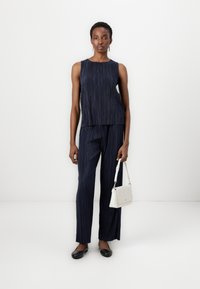 Navy two-piece outfit with pleated texture; sleeveless top and loose pants. Model holds a small white handbag, wearing black flats.
