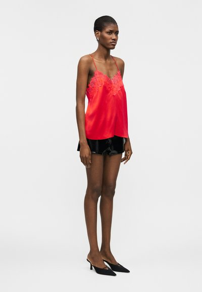 Γυναίκα που φοράει κόκκινο σατέν καμιζόλ με δαντελένιο τελείωμα, μαύρα σατέν σορτς και μαύρες γόβες με μυτερή μύτη, στέκεται μπροστά σε ένα απλό ανοιχτόχρωμο φόντο.