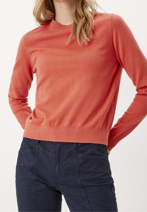 Femme portant un pull en maille corail à manches longues et un pantalon bleu marine foncé, les mains posées sur les hanches, sur un fond clair uni.