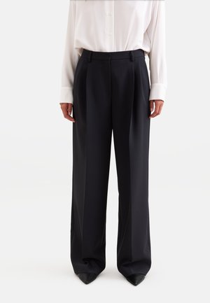 Vrouw draagt zwarte broek met wijde pijpen en een witte blouse met lange mouwen en knopen, staand met handen ontspannen langs het lichaam.