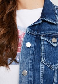 Denim jacket featuring silver snap buttons, structured collar, and blue wash. Worn over a white T-shirt with a graphic print.
