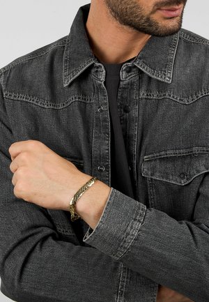 Giacca di denim grigia scura con chiusura a bottoni, due tasche sul petto e un bracciale a catena dorato al polso, che mette in mostra una finitura metallica lucida.