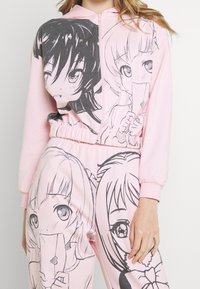 Completo con felpa e pantaloni rosa caratterizzato da illustrazioni grafiche nere di due personaggi. Il tessuto è morbido e presenta una vita e polsini elasticizzati.