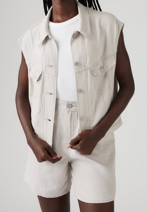 Personne portant un gilet en denim sans manches beige clair, un short taille haute assorti et un haut blanc à manches courtes côtelé, les mains ajustant le devant du gilet.