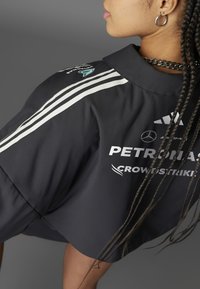 Tamna, skraćena jakna s bijelim prugama, sa logotipovima: Petronas, CrowdStrike i Adidas. Glatki materijal s dizajnom s ovratnikom.