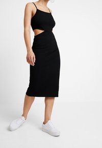 Schwarzes, figurbetontes Midikleid mit seitlichen Ausschnitten, dünnen Trägern und glatter Textur. Kombiniert mit weißen Sneakers für einen Kontrast. Einfaches Design, ohne Muster.