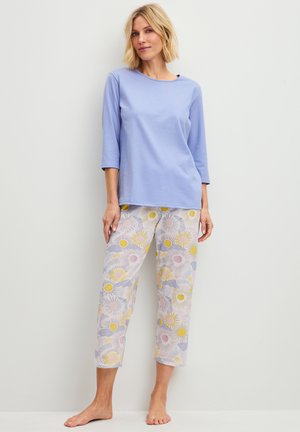 Vrouw met kort blond haar, draagt een lichtblauw driekwart mouw topje en gebloemde cropped broek, staat blootsvoets tegen een witte muur.