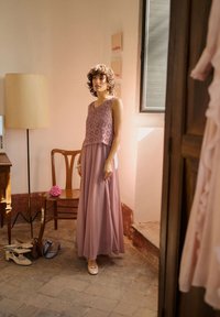 Femme en robe longue mauve et haut en crochet, debout près d'une chaise en bois avec une fleur rose, à côté d'une lampe sur pied et de chaussures sur un sol carrelé.