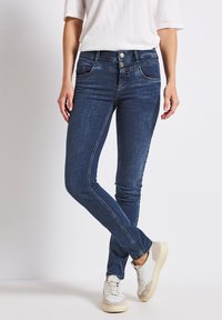 Jean skinny bleu avec une conception à cinq poches, fermeture à bouton métallique et zip, légères décolorations, et tissu en denim doux, accompagnés de baskets blanches.
