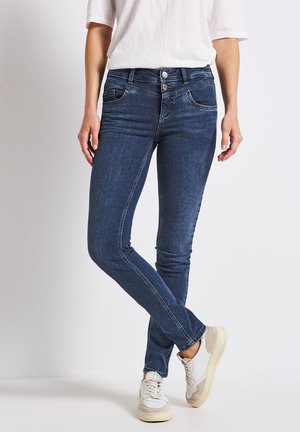 Jean skinny bleu avec une conception à cinq poches, fermeture à bouton métallique et zip, légères décolorations, et tissu en denim doux, accompagnés de baskets blanches.