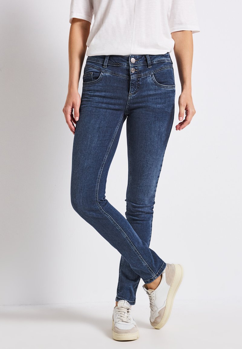 Jean skinny bleu avec une conception à cinq poches, fermeture à bouton métallique et zip, légères décolorations, et tissu en denim doux, accompagnés de baskets blanches.