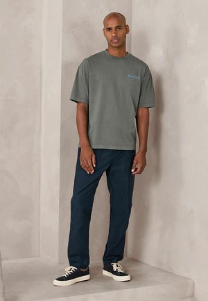 Mann steht vor einer beigen Wand und trägt ein graues Pink Floyd T-Shirt, marineblaue Hose und schwarze Sneakers mit weißen Schnürsenkeln und Sohlen.