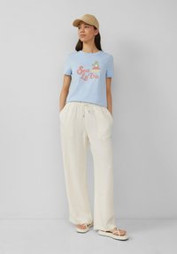 Lichtblauwe t-shirt met de opdruk "Sea La Vie", gecombineerd met losse witte linnen broek en beige sandalen, afgemaakt met een beige pet.