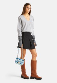 Šedý svetr s V-výstřihem a žebrovanými konci rukávů, černá skládací sukně, hnědé holínky do výšky kolen a modro-bílá geometrická kabelka.