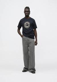 Mees, kes seisab mustas tenniseklubi t-särgis, laienevate hallide teksapükstega ja mustade kingadega, valge ühetoonilise tausta ees.