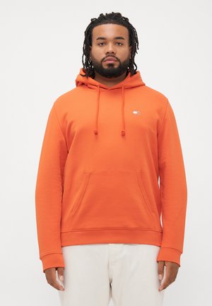 Mand med skæg og dreadlocks iført en orange hættebluse med frontlomme og cremefarvede bukser, stående mod en hvid baggrund.
