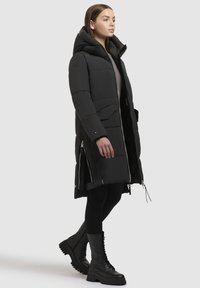Schwarze Pufferjacke mit Kapuze, ausgestattet mit einem Reißverschluss vorne, seitlichen Reißverschlüssen und großen Taschen. Glatte Textur und knielanger Schnitt. Schwarze Stiefel getragen.