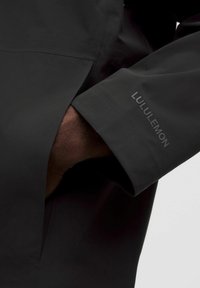 Čierna nepremokavá bunda s hladkou textúrou, viditeľnými manžetami a prešitými detailmi. Obsahuje logo Lululemon v kontrastnej šedej farbe.