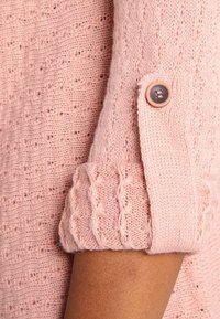 Rosa gestrickter Pullover mit einem strukturierten Muster, gerippten Bündchen mit einem Zopfmuster und einem Knopfdetail am Ärmel.