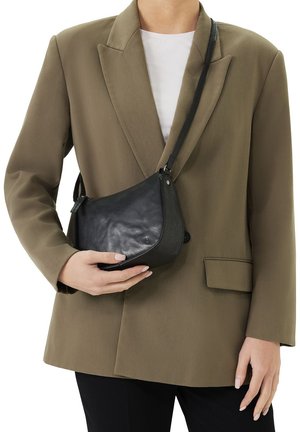 Zwarte leren schoudertas met een gebogen vorm, verstelbare schouderband en subtiele logo-detail, vastgehouden tegen een blazer in neutrale tinten.