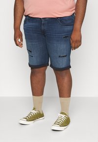 Pantalones cortos de mezclilla en azul oscuro con parches desgastados, puños enrollados, combinados con zapatillas verdes y calcetines beige. El cuerpo se presenta de manera casual.