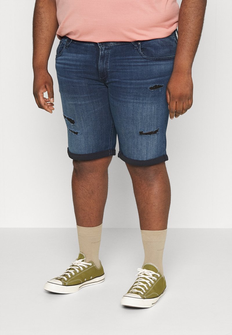 Pantalones cortos de mezclilla en azul oscuro con parches desgastados, puños enrollados, combinados con zapatillas verdes y calcetines beige. El cuerpo se presenta de manera casual.