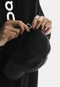 Căciulă neagră cu o visieră curbată, ajustată de mâini. Căciula are o curea albă ajustabilă și o textură din material.