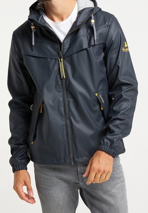 Man draagt een marineblauwe regenjas met rits en capuchon en gele ritslipjes, gecombineerd met lichtgrijze jeans en een wit overhemd.