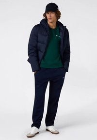 Granatowa kurtka puchowa, zielony sweter, granatowe spodnie dresowe i białe sneakersy. Nosi czarną czapkę. Kurtka puchowa ma przeszywane panele i kaptur.