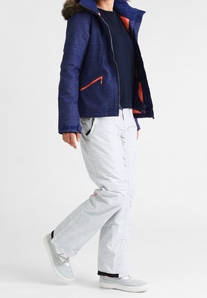 Marineblauw jack met bont gevoerde capuchon, oranje accenten, gecombineerd met lichtgrijze broek. De broek heeft een gestructureerd ontwerp en zakken. Witte schoenen.