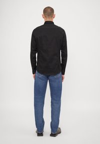 Svart skjorta med knappar, med en enkel struktur, krage och långa ärmar, kombinerad med blå jeans och grå skor, sedd bakifrån.