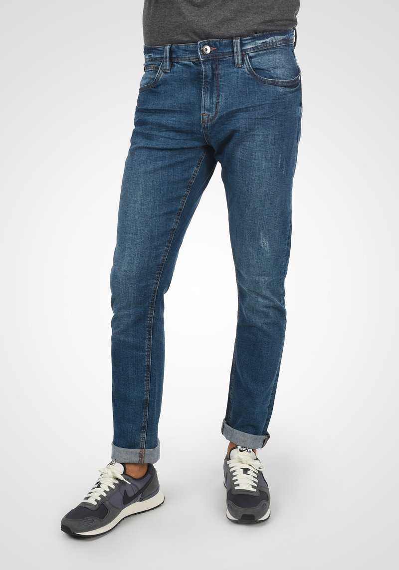 Kék farmernadrág, slim fit szabásban, felgörbített szárakkal, enyhe kopással és klasszikus öt zsebes kialakítással. Szürke sneaker cipővel párosítva.