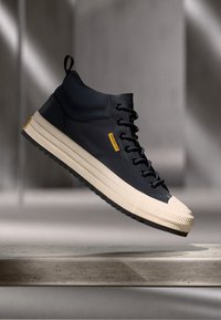Des baskets montantes en matériau bleu marine avec une semelle en caoutchouc crème, dotées de lacets noirs et d'un logo jaune sur le côté.