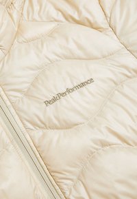 Veste rembourrée couleur crème avec un design matelassé, finition brillante et logo "PeakPerformance" subtil en doré. Dispose d'une fermeture éclair.