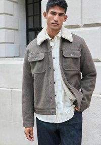 Braune Wollsjacke mit einem Lammfellkragen, Druckknöpfen und zwei Brusttaschen. Über einem karrierten Hemd und dunklen Jeans getragen.