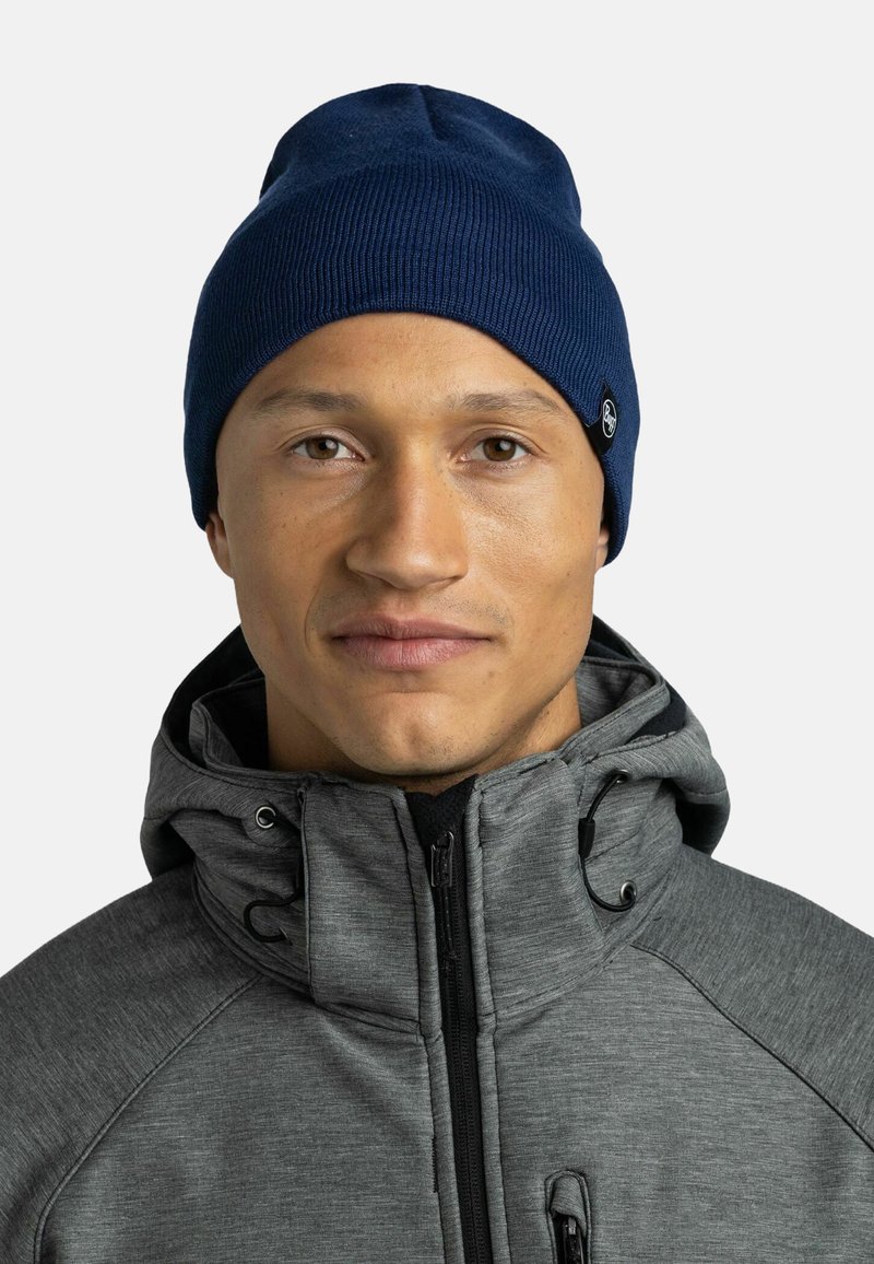 Navy knitted beanie with a ribbed texture. Worn with a grey zip-up jacket featuring a high collar and adjustable drawstrings.