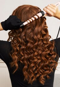 Lockenstab mit pinkem Barrel und schwarzen Akzenten. Das Haar ist wellig und rot-braun, zu engen Locken frisiert. Eine handschuhtragende Hand hält den Lockenstab.