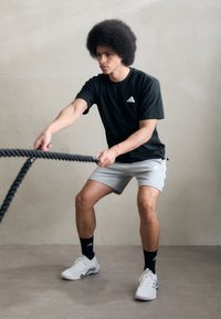 Mustus lühikeste varrukatega T-särk, hallid lühikesed püksid, valged spordijalatsid. Inimene teostab harjutust lahingunööridega minimalistisch keskkonnas.