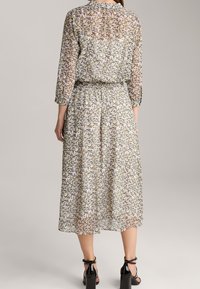 Długa, kwiatowa sukienka z przezroczystej tkaniny, zdobiona wielokolorowym wzorem małych kwiatków, z elastycznym pasem i rękawami o długości trzech czwartek. Widać czarne obcasy.