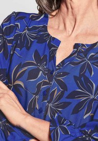 Blusa floreale blu con scollatura a V, dettagli con bottoni e un tessuto testurizzato con motivi di foglie blu scuro e marrone chiaro.