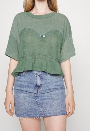 Femme portant un crop top vert ample orné de dentelle, associé à une mini-jupe en denim bleu clair à ourlet effiloché.