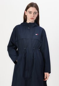 Impermeabile blu navy realizzato in materiale leggero, con cappuccio, cordino in vita e tasche laterali. Dettaglio del logo sul petto.